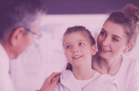 A little girl is sitting on her mother’s lap at a doctor’s appointment. The little girl is smiling whilst the doctor comforts her by putting his arm on her shoulder